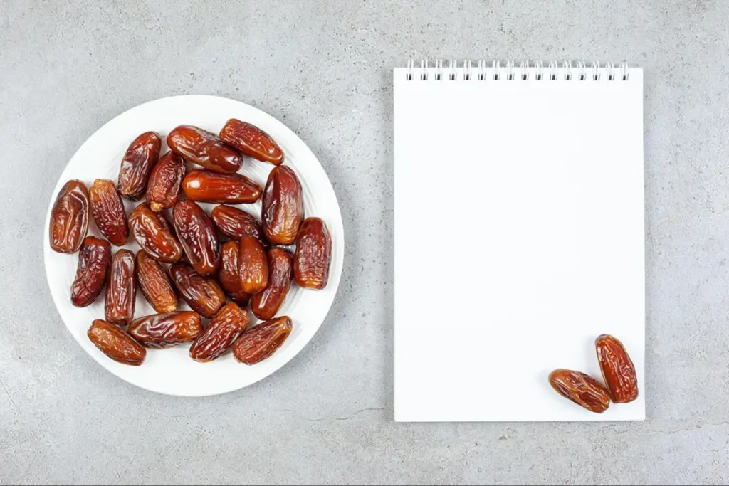A white plate full of premium dates next to a blank notebook, symbolizing the planning of a science-backed nutritional profile and macro intake for athletes.